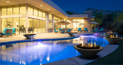 Luxurious modern home with a glowing pool, surrounded by multiple fire water bowls, and outdoor seating under the night sky.