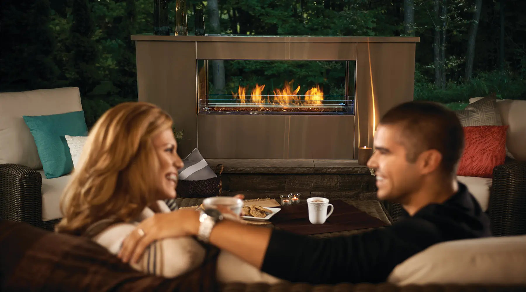 Couple enjoying a cozy evening by a modern see-through outdoor gas fireplace by Summit Fire Designs, sharing warm drinks.